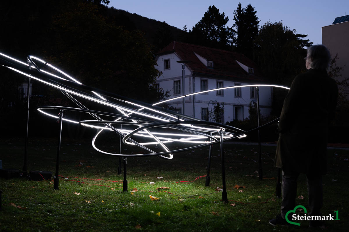 Eine Person betrachtet Miriam Hamanns „Planet 9“ mit verschlungenen weißen Lichtbahnen im Schlosspark Eggenberg.