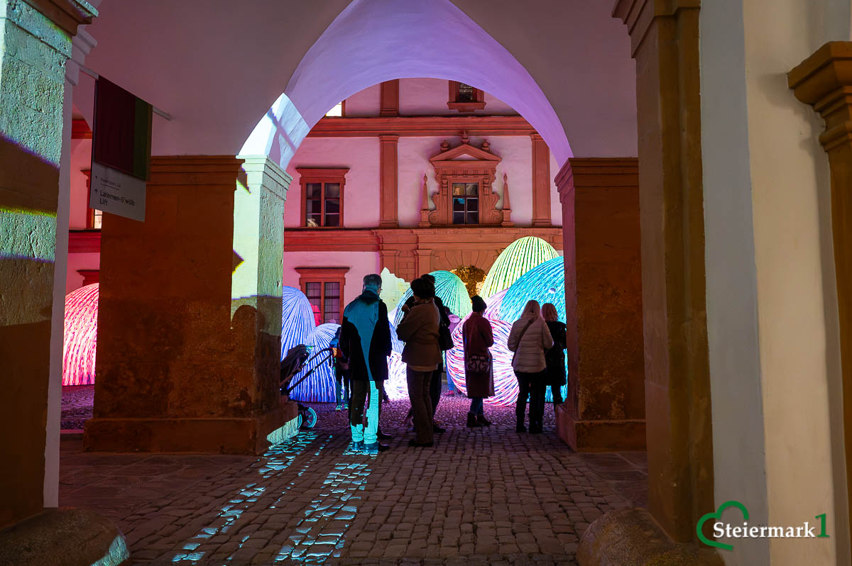 Blick durch einen Torbogen auf Gäste und die bunt leuchtenden „Whispering Mountains“ von ENESS in Schloss Eggenberg.